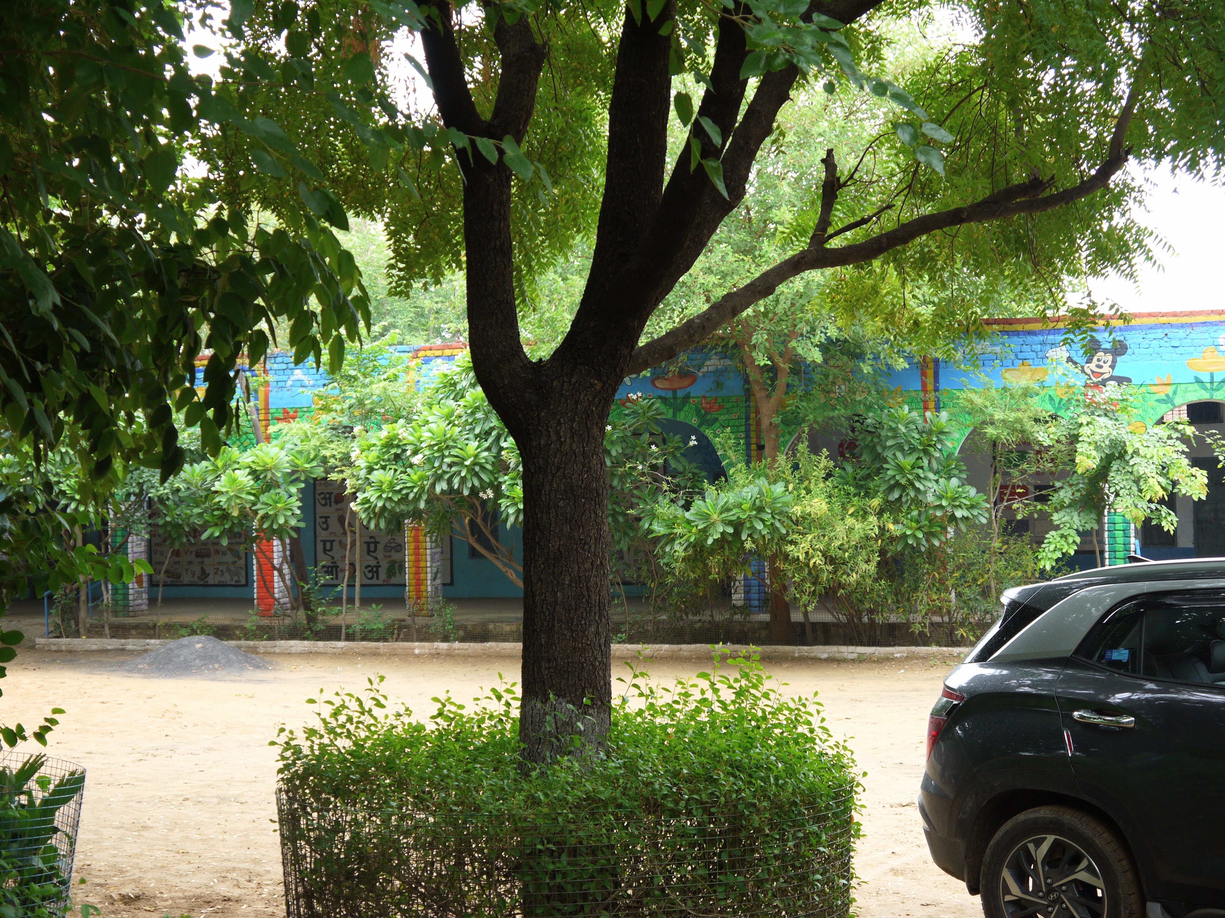 Lush trees surround an open courtyard enclosed by a brick school painted in colourful educational murals.