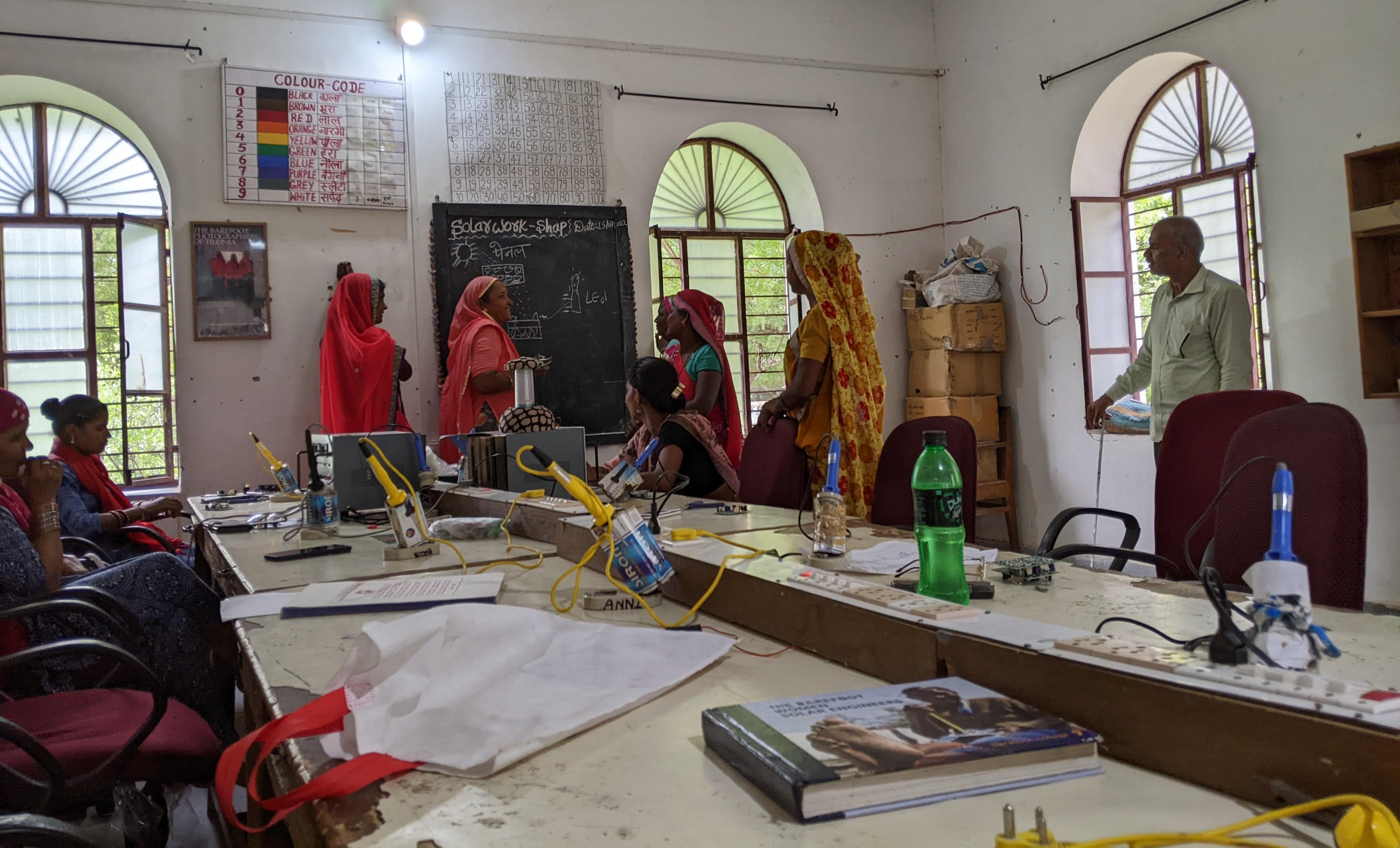 Eight women in traditional Rajasthani clothing gather around a long worktable with soldering irons and various other tools on it. A chalkboard and a series of posters on one wall describes lessons on electrical design.